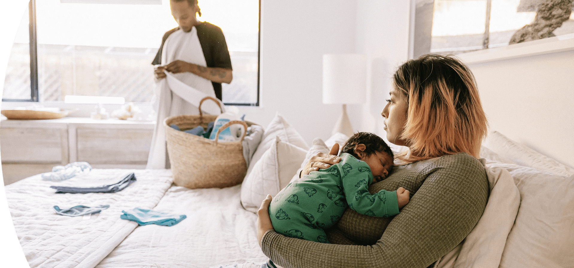 A woman holding a newborn while her partner folds laundry in the background